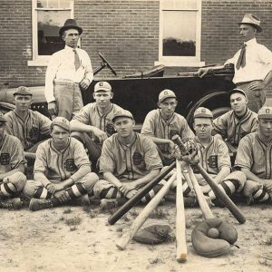 Group of men dressed in baseball uniforms seated on ground with two men in ties standing behind them