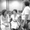 Two white women sitting and one white woman standing wearing skirts and dresses and talking in room with rows of chairs