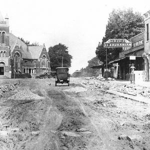 Old fashioned car driving down dirt road between rows of multistory buildings