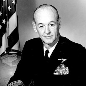 Older white man with in military uniform with American flag and a globe in the background