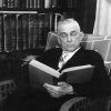 Portrait of a man in formal attire reading in a wicker chair next to a bookcase.