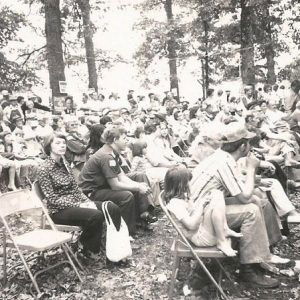 Crowd of white men women and children sitting in folding chairs under trees at political rally