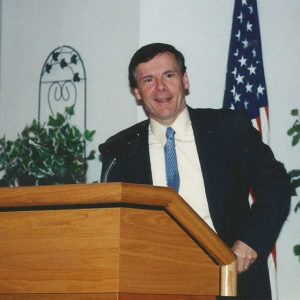 White man in suit and tie speaking at lectern