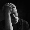 Old white man with beard sitting with his right hand on his head