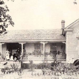 White family on steps of single-story house with covered porch and fence and man with horse nearby