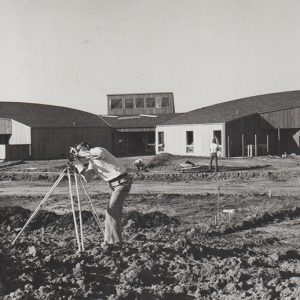 White man with surveying equipment standing in field with white men and building with rounded wings and skylight behind them