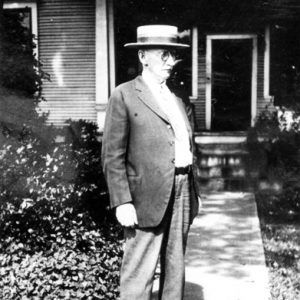 white man in straw hat stands outside wooden house