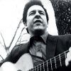 White man in button up shirt and jacket playing acoustic guitar in front of tree trunk