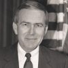 White man smiling in suit and tie with flag behind him