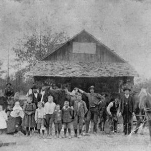 Group of white men women children and horse drawn wagon outside single-story building