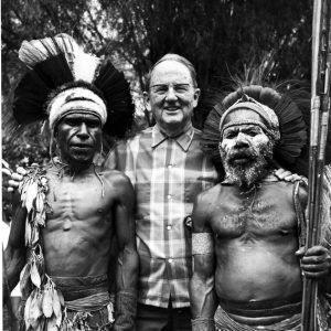 older white man in flannel shirt and glasses with his arms around two dark-skinned men in headdresses holding spears