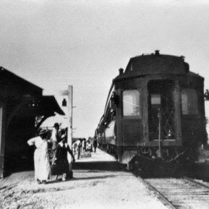 Men and women waiting on depot platform with train on tracks