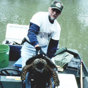 Gloved white man on boat exhibiting large gaping alligator snapping turtle among coolers toolbox bucket