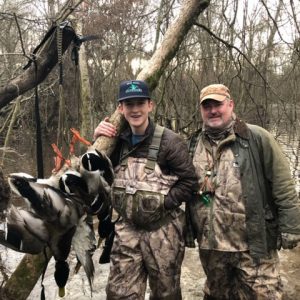 Young white teenage boy and older white man in hunting gear in swamp with ducks hanging from a tree