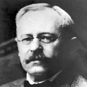 White man with small round glasses and mustache in suit and bow tie