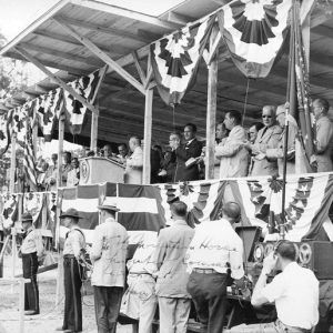 White man at lectern speaking to white crowd from a covered stage decorated with bunting