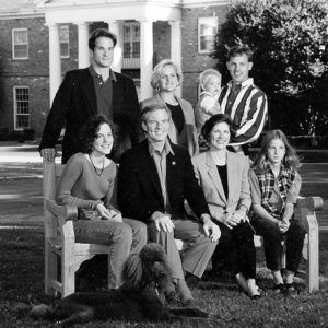 White family posing on bench with dog on lawn in front of multistory brick house with covered porch