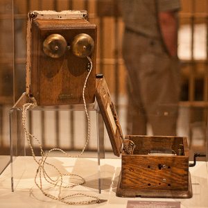 Telephone bells and open wooden box with crank handle in glass display case