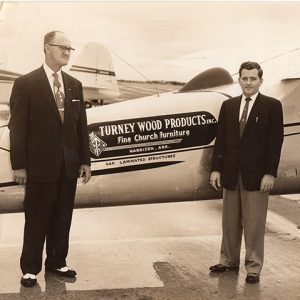 Two white men in suit and tie posing with "Turney Wood Products" logo on airplane