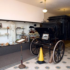 Black stagecoach with wooden wheels in museum gallery next to glass display case