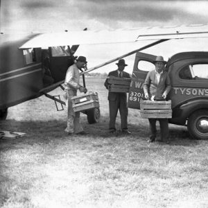 Three white men transferring crates from an airplane to a car that says "Tyson's Hatchery" on the side