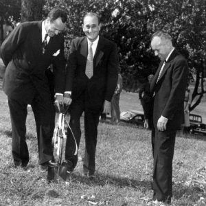 Three white men in suits laughing while using a shovel to break ground with tree and spectators behind them