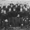 Group of young white men in matching dark colored uniforms standing together on field