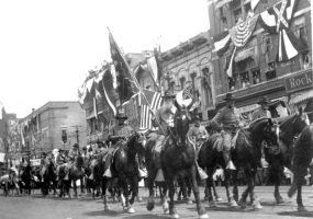 White men in Confederate uniforms and hats holding Confederate flags ride on horseback in a parade