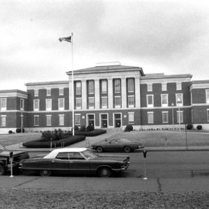 Multistory brick courthouse with six columns and grounds, with street and parked cars in foreground