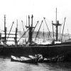 Ship in harbor with lifeboats floating nearby and shore in the background