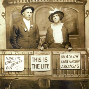 White man and woman in suit and dress posing with fake train car set featuring signs such as "I love the cows and chickens but" and "This is the life" and "On a slow train through Arkansas."