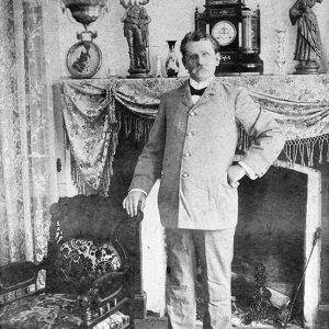 White man with mustache in suit standing in room with fireplace