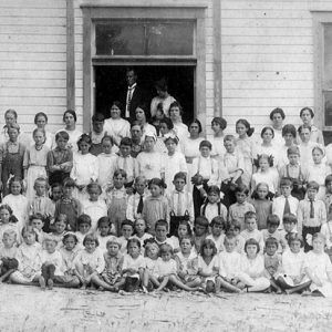 Large group of adults and children posing in front of wooden building