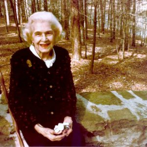 older white woman in cardigan on stone wall in front of woods