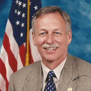 White man with mustache smiling in suit and tie with flag behind him