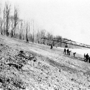 Crane barge with arm extended over shore from river with group of workers digging below it