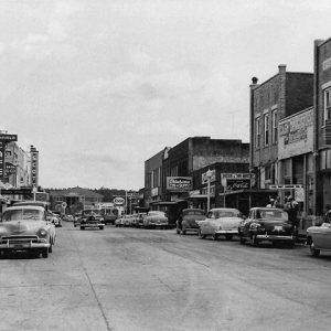 Parked cars on street with multistory brick storefronts and signs on both sides