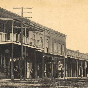 People standing on covered sidewalk outside multistory storefronts with balcony on dirt street