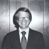 White man in suit and glasses standing in front of wood paneling