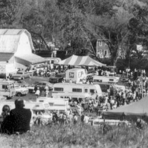 Recreational vehicles parked near a large barn with people around them and a large tent behind a two-story house