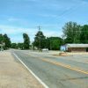 Single-story buildings and parking lots on either side of two-lane road with trees in the background