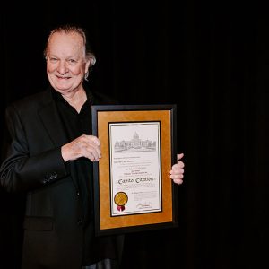 White man wearing black holding framed "Capitol Citation" document