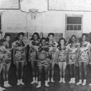 African-American girls in matching uniforms with African-American men in suits standing below basketball hoop