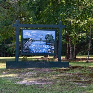 Bird and lake on "White Oak Lake State Park" sign in flower bed with trees behind it