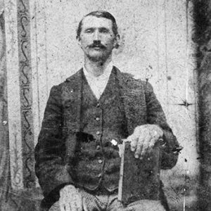 White man with mustache wearing a suit sitting with book on this knee