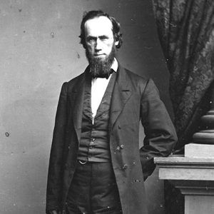 White man with beard standing in suit with his arm behind his back