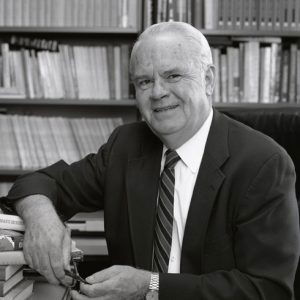 Older white man smiling in suit and tie sitting at desk with bookshelves behind him