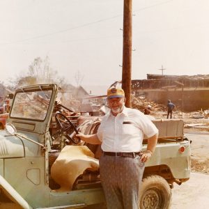 Older white man with hat standing next to Jeep at site of damaged house