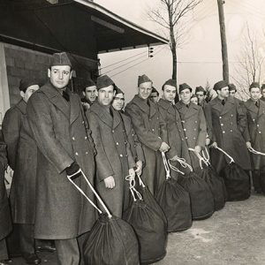 Group of young white men in military coats and caps standing with duffel bags