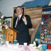 White man in suit speaking at lectern with flowers in the foreground